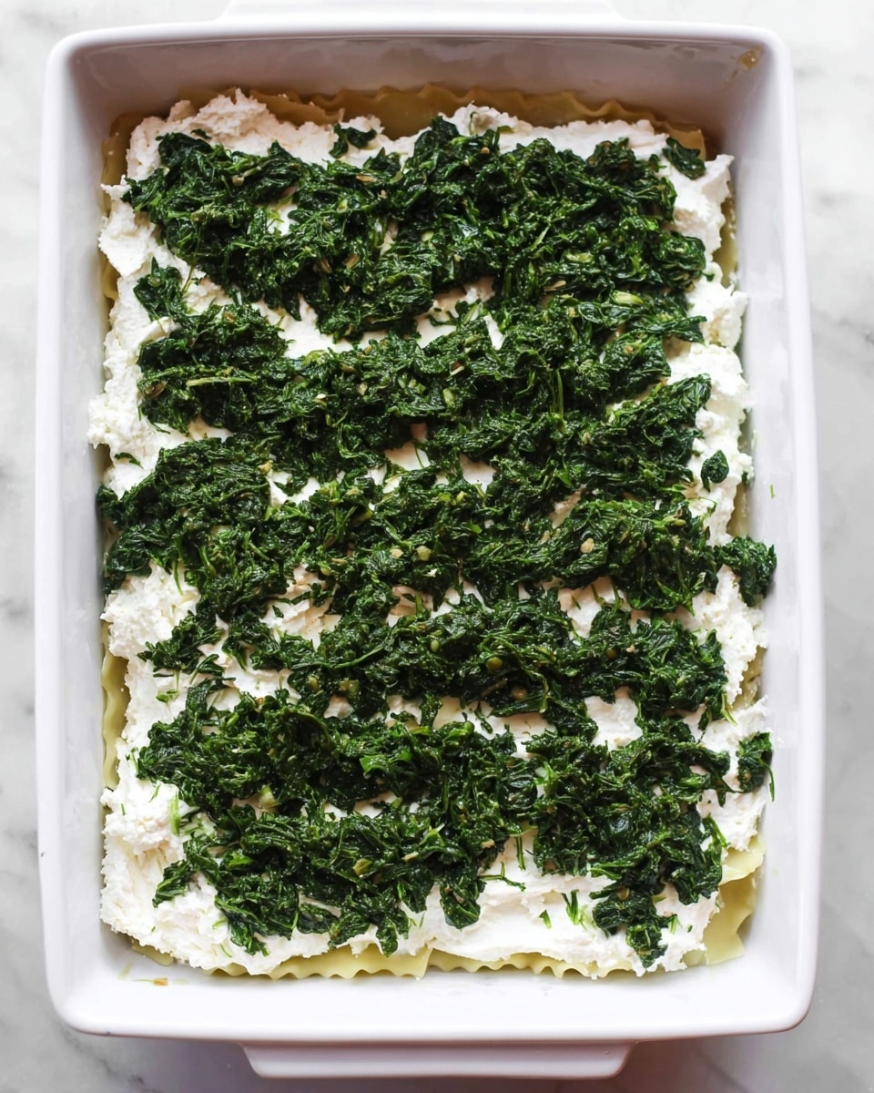 The image shows a white rectangular baking dish with a layer of lasagna noodles at the bottom visible around the edges. On top of the noodles is a thick layer of white ricotta cheese spread evenly, with a slightly rough texture. Covering the cheese is a generous layer of dark green cooked spinach, roughly chopped and spread out, creating a dense topping. The dish is set on a white marbled surface. photo taken with an iphone --ar 4:5 --v 7