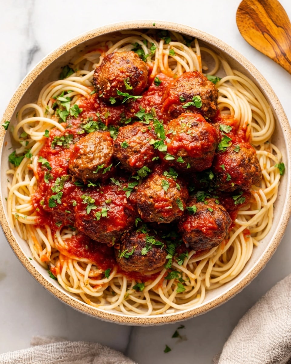 A round bowl filled with a base layer of light tan spaghetti noodles that are loosely arranged in circles. On top of the noodles, there is a layer of deep red tomato sauce that lightly covers parts of the pasta. The top layer consists of several brown meatballs glazed with tomato sauce, scattered evenly around the bowl. Small pieces of green herbs are sprinkled over the meatballs, sauce, and noodles, adding a fresh touch. The bowl rests on a white marbled surface, with part of a wooden utensil visible on the right side. photo taken with an iphone --ar 4:5 --v 7