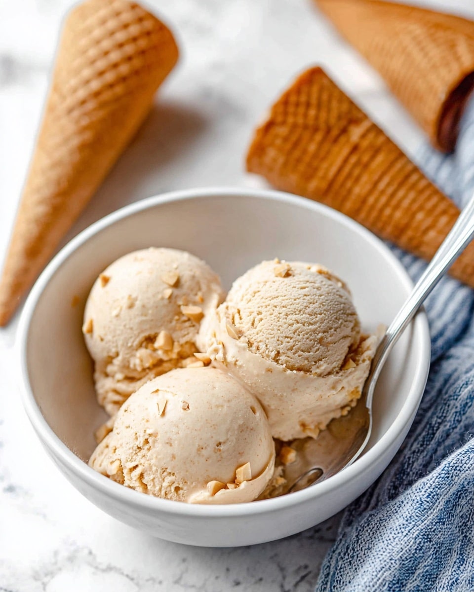 A white bowl holds three round scoops of light brown ice cream with a smooth texture and small nutty bits visible throughout. A silver spoon rests inside the bowl on the right side. In the background, there are two ice cream cones lying on a white marbled surface, which has soft, subtle gray veins. A blue and white striped cloth is partially under the bowl and extends to the right side of the image. The scene is bright and clear with a close-up focus on the ice cream scoops. photo taken with an iphone --ar 4:5 --v 7