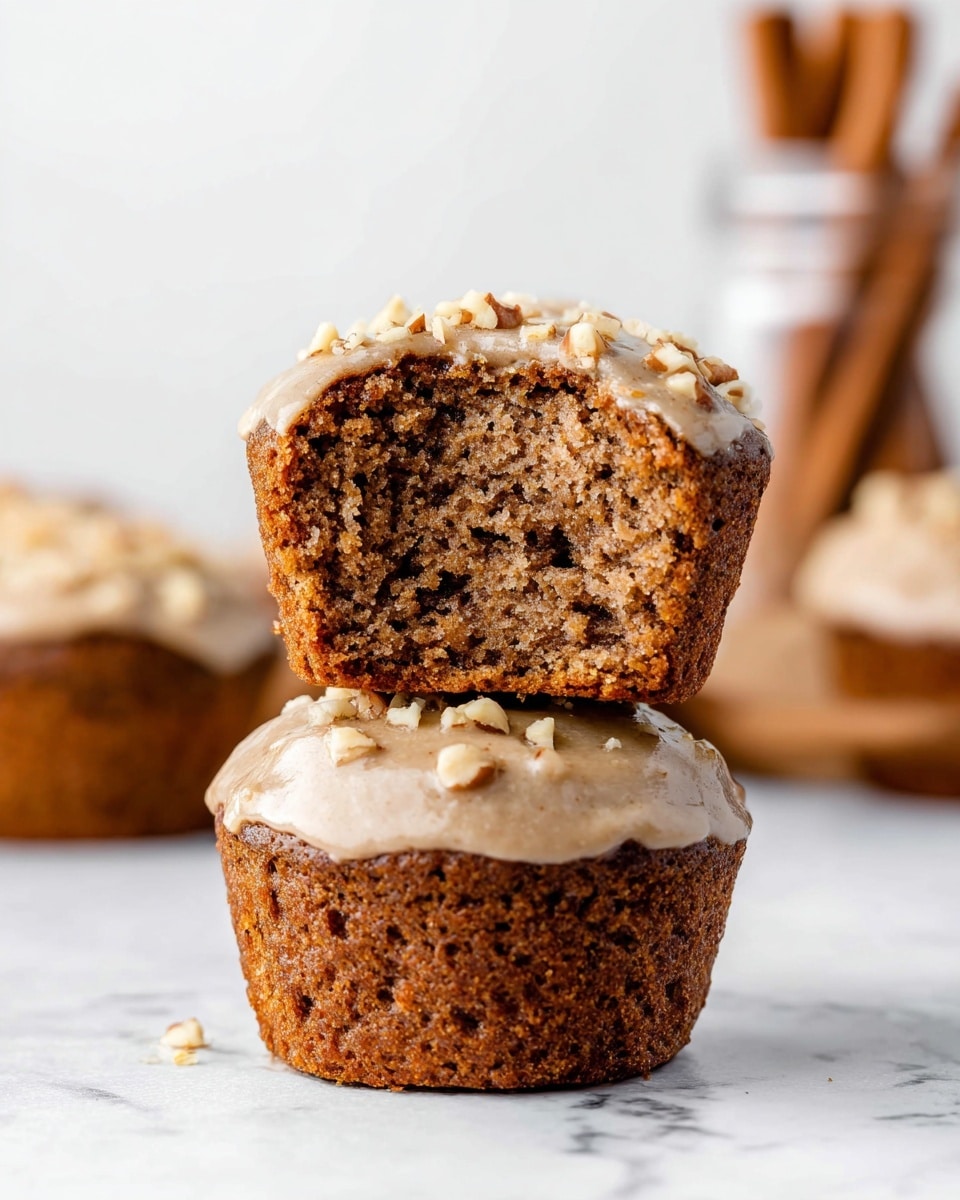 Two brown muffins are stacked on a white marbled surface. The bottom muffin is whole with a light beige icing topped with small nut pieces covering the top. The top muffin is cut in half, showing its soft, moist, and dense brown inside, with the same light beige icing and nut pieces on top. In the background, blurred muffins and a jar with cinnamon sticks add depth to the image. Photo taken with an iphone --ar 4:5 --v 7