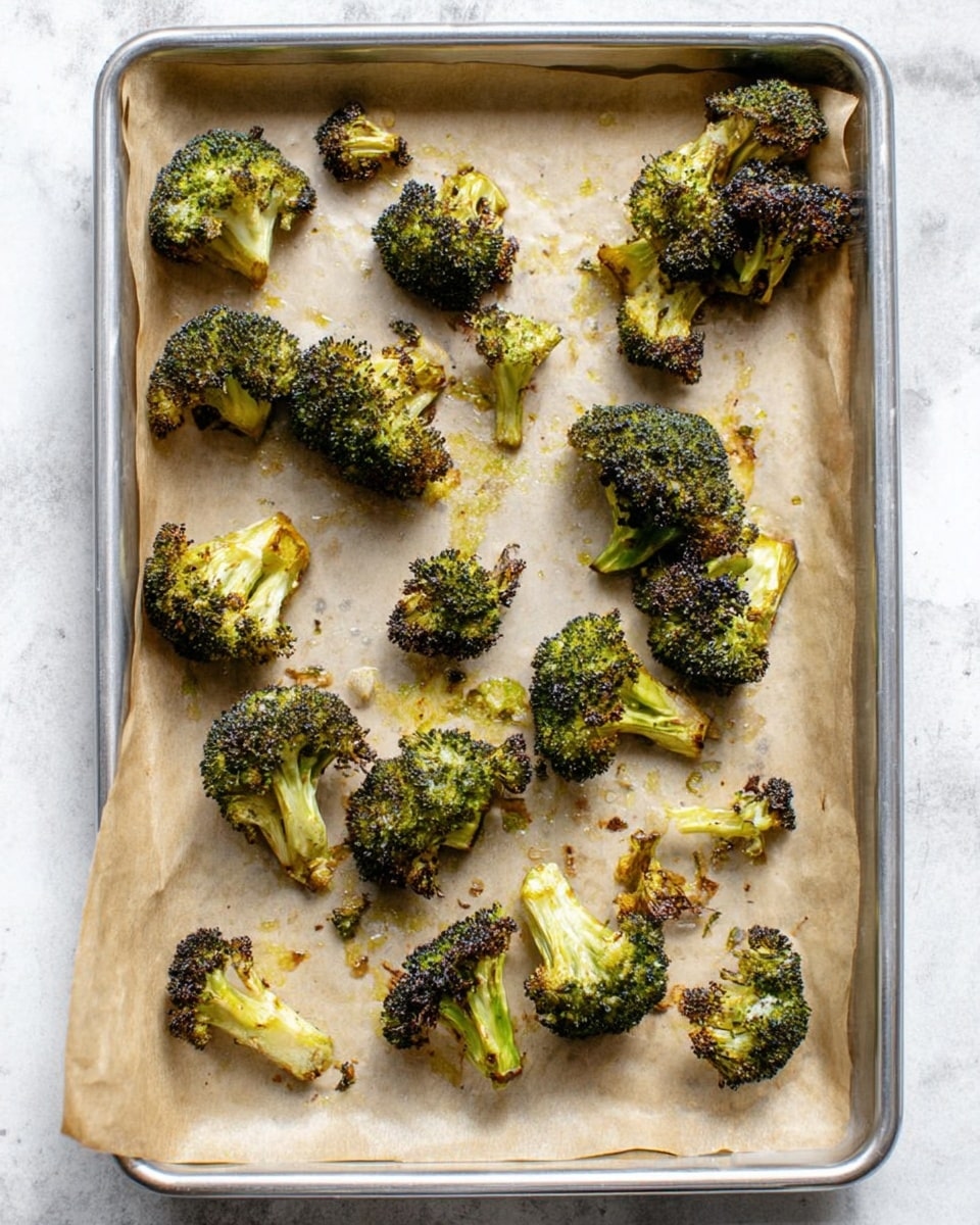 The image shows a silver baking tray lined with light brown parchment paper on a white marbled surface. Scattered across the tray are about twenty small roasted broccoli florets, each with a mix of deep green and slightly charred dark brown areas on the tops, while the stems are a pale green to light yellow color. The florets vary in size and have a crispy texture, with some showing small bits of charred edges. The tray and lighting give a homey, fresh roasted vegetable look. Photo taken with an iphone --ar 4:5 --v 7