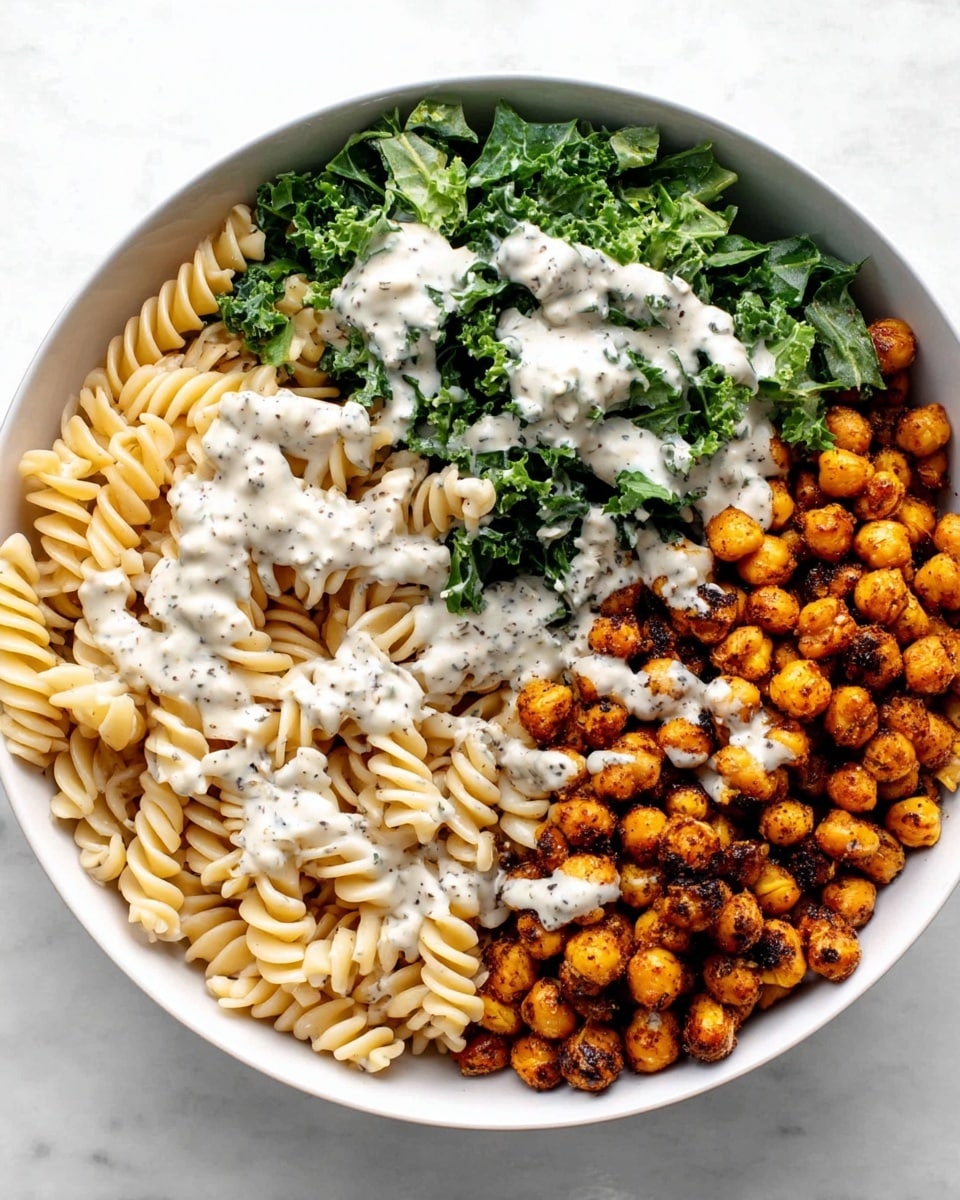 The image shows a white bowl filled with a three-part dish arranged side by side. On the left side, there are light beige spiral pasta pieces. The middle section has dark green chopped leafy kale. On the right side, there are dark orange roasted chickpeas. A light creamy white sauce with black specks is drizzled evenly over all three sections, creating a contrast with the colors underneath. The bowl is placed on a white marbled surface photo taken with an iphone --ar 4:5 --v 7