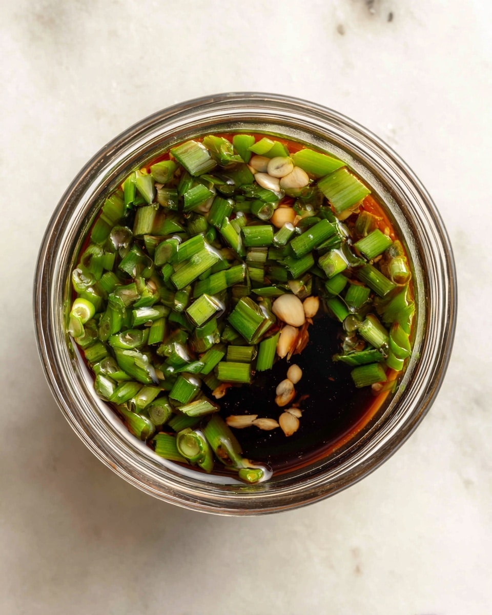 A top view of a small clear glass bowl filled with a dark soy sauce mixture, layered with chopped green onions scattered evenly on the top, giving a fresh green contrast to the deep brown liquid beneath. Small bits of garlic or ginger float just under the surface, adding light tan spots near the center. The sauce is shiny, showing light reflections, and the bowl sits on a white marbled surface. photo taken with an iphone --ar 4:5 --v 7