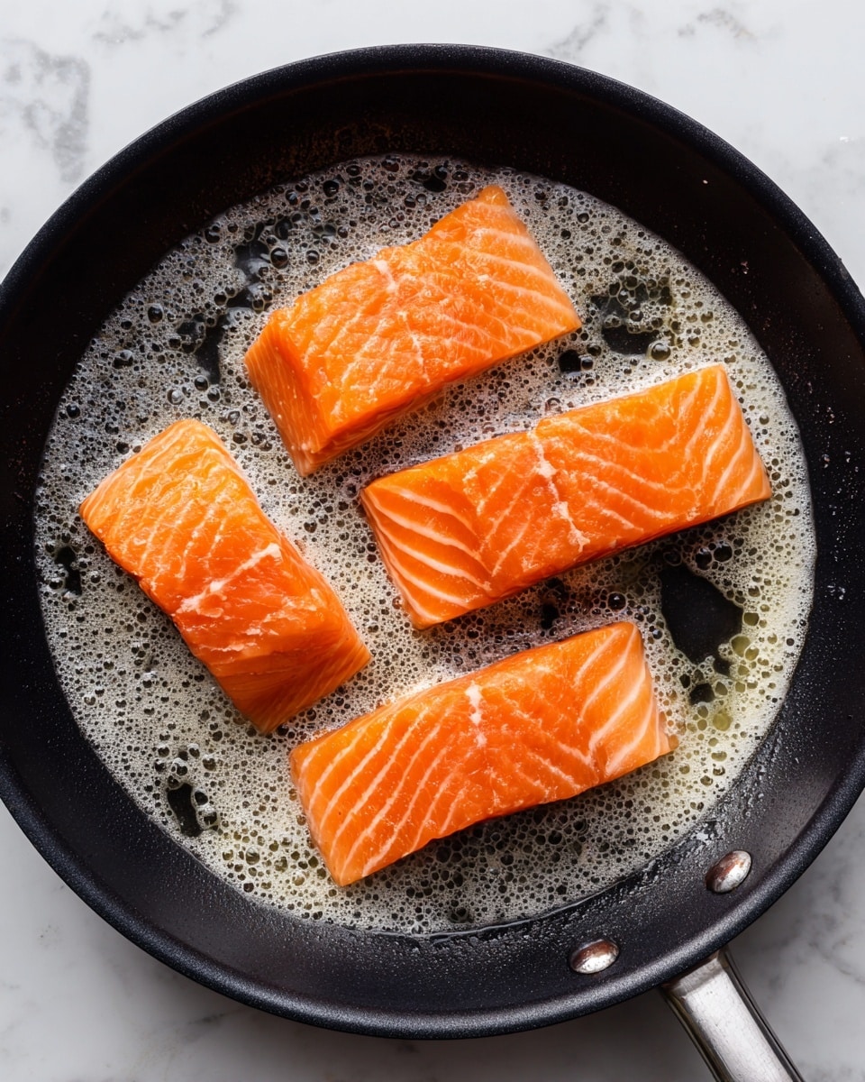 A black frying pan sits on a white marbled surface, holding three pieces of raw salmon fillets. The salmon pieces are bright orange with visible lines of fat and a slightly shiny texture. They are evenly spaced in the pan, sizzling in melted butter that bubbles around them. The butter foams white with tiny bubbles, covering part of the pan's bottom. The background is a clean white marbled texture. photo taken with an iphone --ar 4:5 --v 7