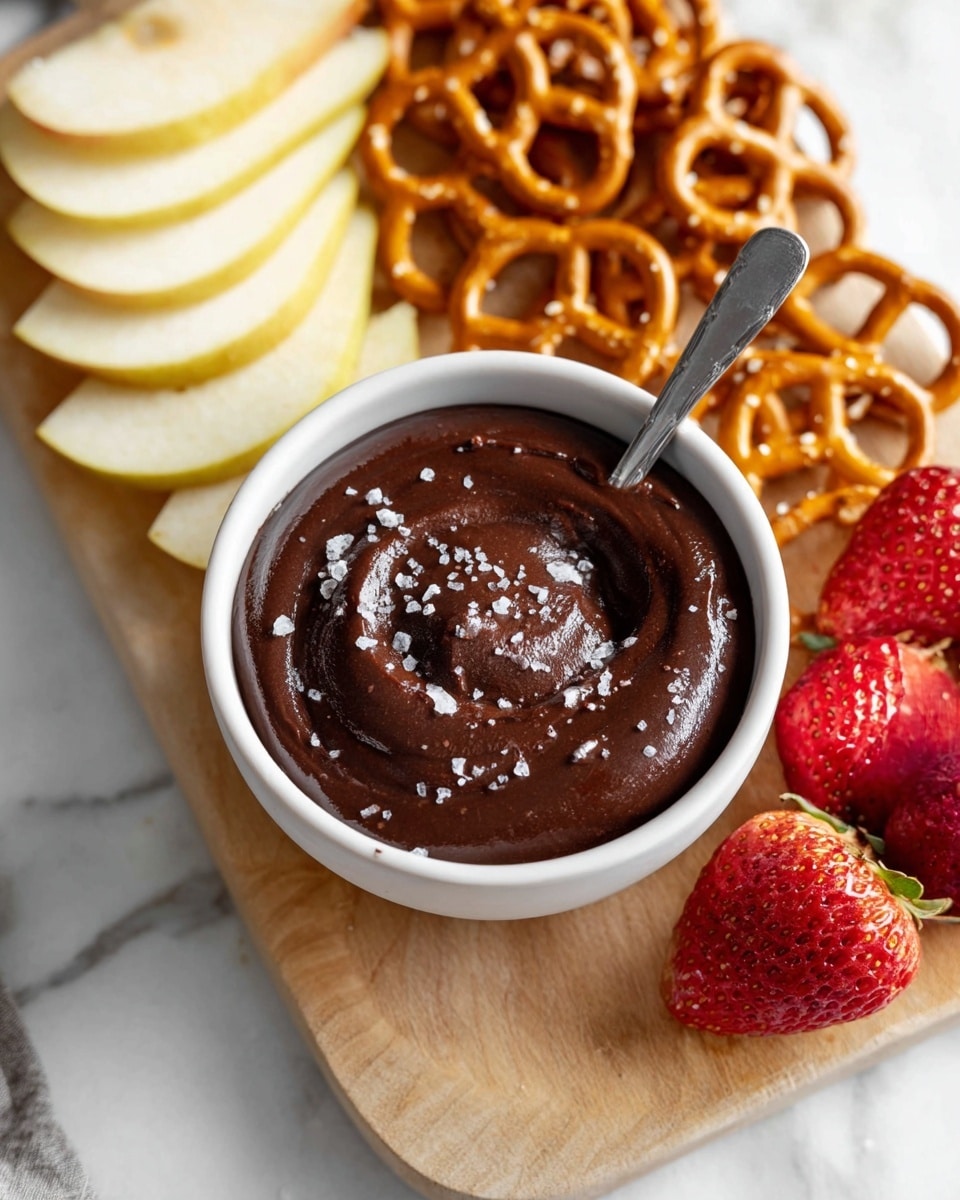 A small white bowl filled with thick, dark brown chocolate dip sprinkled with coarse sea salt on top, a silver spoon resting inside the dip. Behind the bowl, there are layered slices of light yellow pear arranged in a small pile on the left, golden brown pretzels clustered on the right, and several red strawberries, some whole and some sliced, positioned in the bottom right area. All items are set on a light wooden board placed on a white marbled surface. Photo taken with an iphone --ar 4:5 --v 7