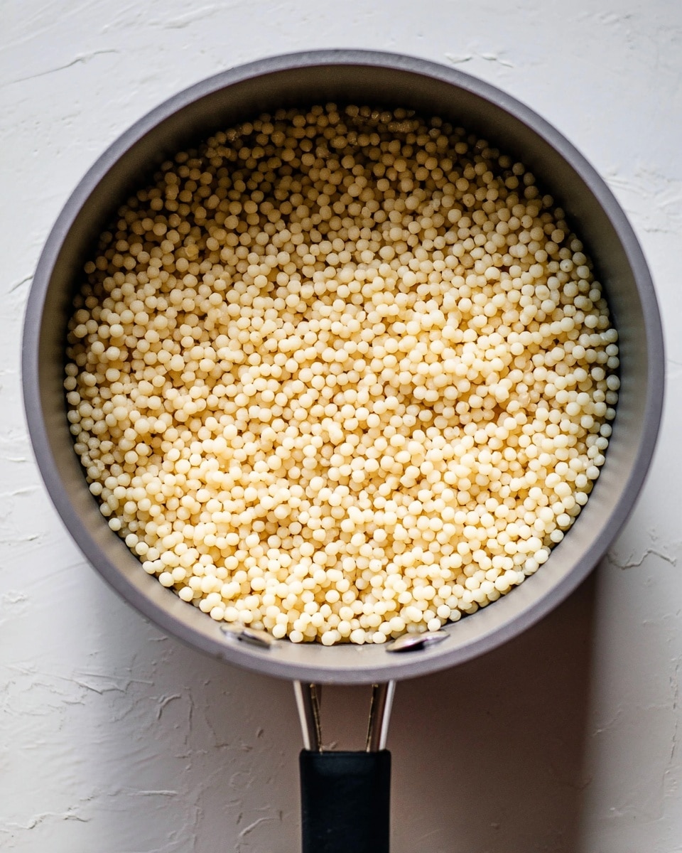 The image shows a single layer of small, round, pale yellow couscous grains filling a gray cooking pot with a black handle. The couscous looks soft and slightly shiny, spread evenly inside the pot. The pot is placed on a white marbled surface, and soft natural light highlights the texture of the grains and the pot's rim. photo taken with an iphone --ar 4:5 --v 7