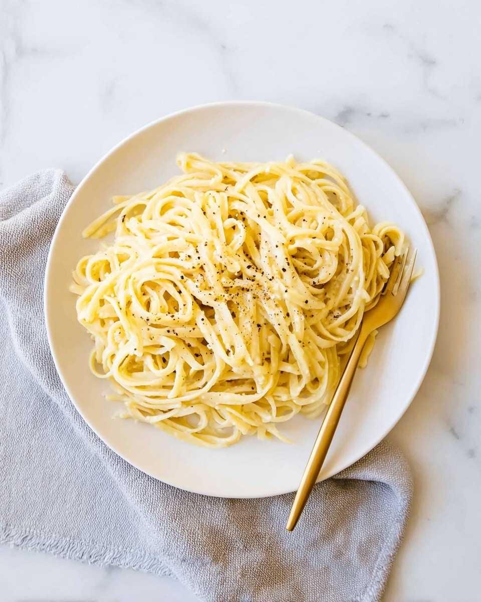 A white plate holds a simple serving of plain yellowish pasta noodles piled loosely in the center, sprinkled lightly with small black pepper flakes. A golden fork is resting on the right side of the plate, slightly submerged in the pasta. The plate sits on a light gray cloth napkin atop a white marbled surface, giving the scene a soft and clean look. photo taken with an iphone --ar 4:5 --v 7