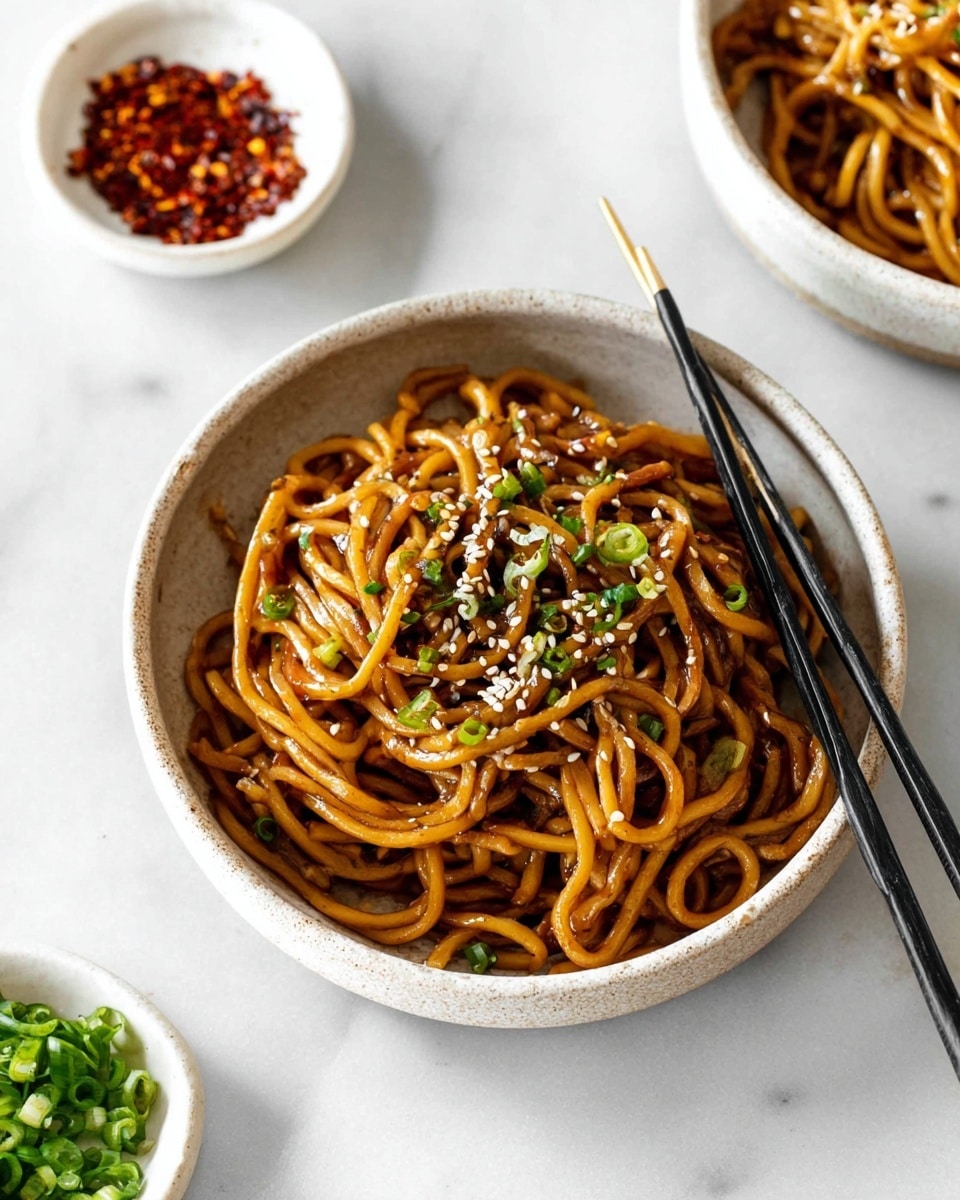 A white speckled bowl is filled with thick brown noodles coated in a glossy sauce, with small pieces of green chives and white sesame seeds sprinkled on top. The noodles are slightly twisted and piled to fill the bowl. Two black chopsticks rest on one side of the bowl. In the background, two small white bowls hold red chili flakes and chopped green chives, all sitting on a white marbled surface. photo taken with an iphone --ar 4:5 --v 7