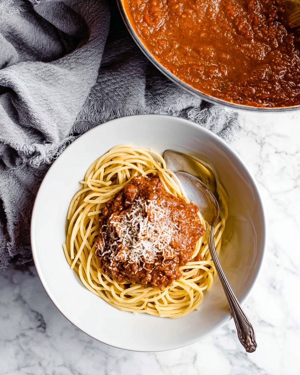 A white bowl filled with a nest of light yellow spaghetti noodles sits on a white marbled surface. On top of the noodles, there is a thick layer of rich red-brown meat sauce, covered with a sprinkling of grated white cheese. A silver fork and spoon rest inside the bowl, leaning against the noodles. To the top left of the bowl, part of a large pot with more red-brown sauce is visible. A soft gray cloth is placed near the bowl on the white marbled surface. photo taken with an iphone --ar 4:5 --v 7