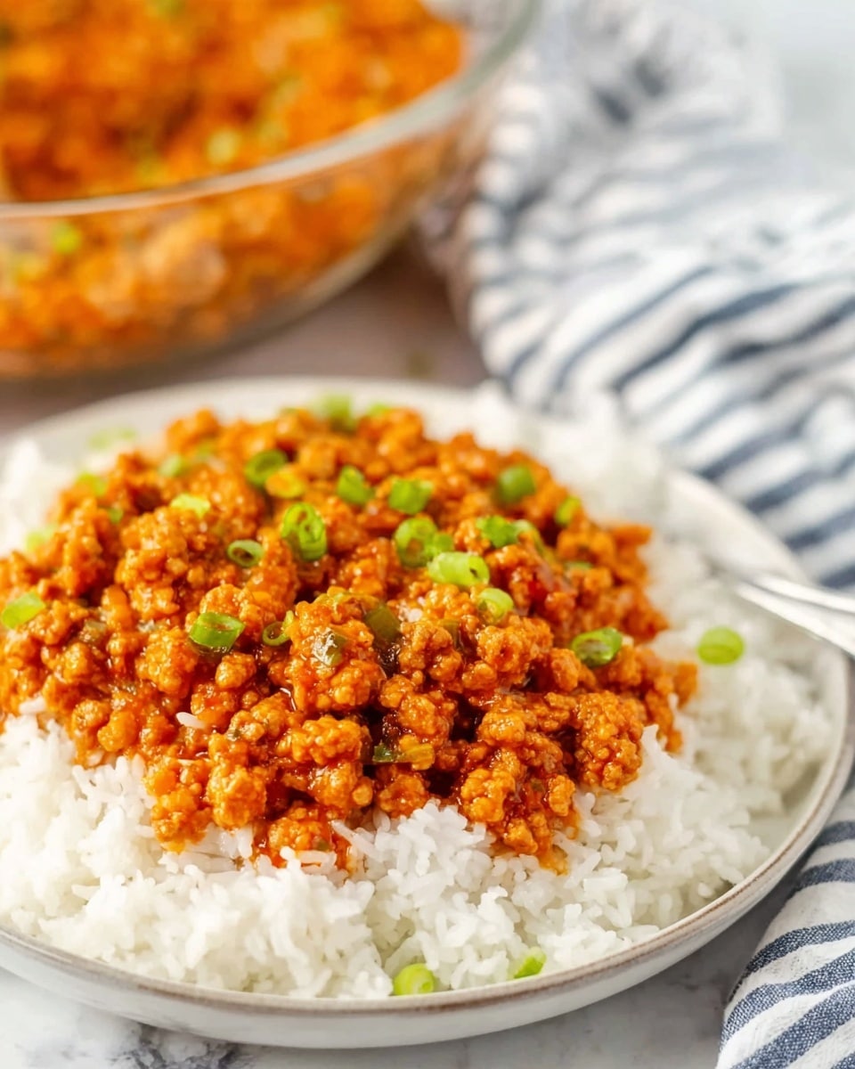 A white plate filled with a base layer of fluffy, white rice, topped with a thick layer of golden-orange saucy ground meat mixed with small bits of vegetables, garnished with small green chopped scallions scattered across the top. In the background, there is a clear glass bowl filled with the same saucy meat mixture resting on a white marbled surface partially covered by a soft cloth with blue and white stripes. The dish looks warm and inviting, with a rich and hearty texture. photo taken with an iphone --ar 4:5 --v 7