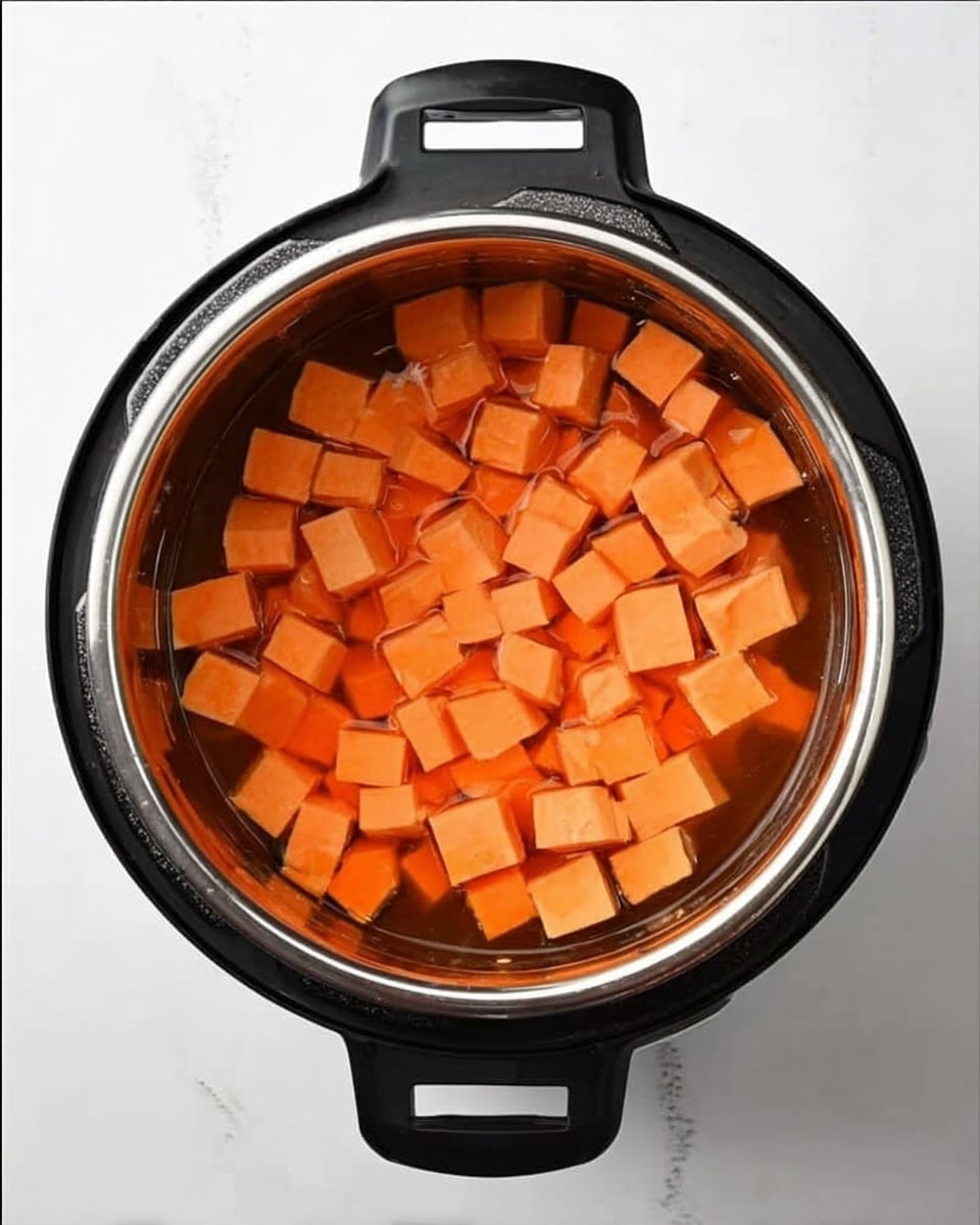 A top view inside an electric pot filled with cubed orange sweet potatoes submerged in water. The sweet potato cubes are evenly spread and cover the whole surface in the pot, which has black handles on both sides. The background shows a white marbled surface. photo taken with an iphone --ar 4:5 --v 7
