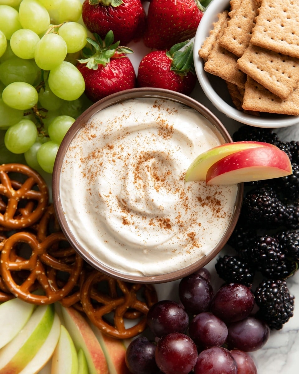 A bowl filled with creamy white dip, sprinkled lightly with brown spice, with a red apple slice dipped into it creating smooth swirls. Around the bowl are layers of fresh fruit and snacks: bright green grapes in a bunch at the top left, red strawberries with green leaves to the left, dark purple grapes at the bottom right, blackberries near the top right, and thin slices of green and red apples arranged near the edges. To the top right of the dip bowl, there is a white bowl filled with light brown rectangular biscuits stacked upright and overlapping. At the bottom left, twisted brown pretzels lie near the fruit on a white marbled surface. Photo taken with an iphone --ar 4:5 --v 7