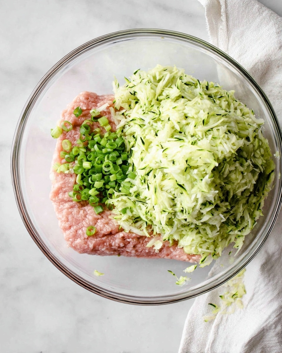 A clear glass bowl sits on a white marbled surface, holding three main layers: at the bottom is a smooth pink layer of raw ground meat, evenly spread. On the left side, finely chopped green onions add small bright green and white pieces over the meat. On the right is a large mound of grated zucchini, pale green with specks of dark green skin, piled high and textured. A white cloth with more grated zucchini rests beside the bowl. Photo taken with an iphone --ar 4:5 --v 7