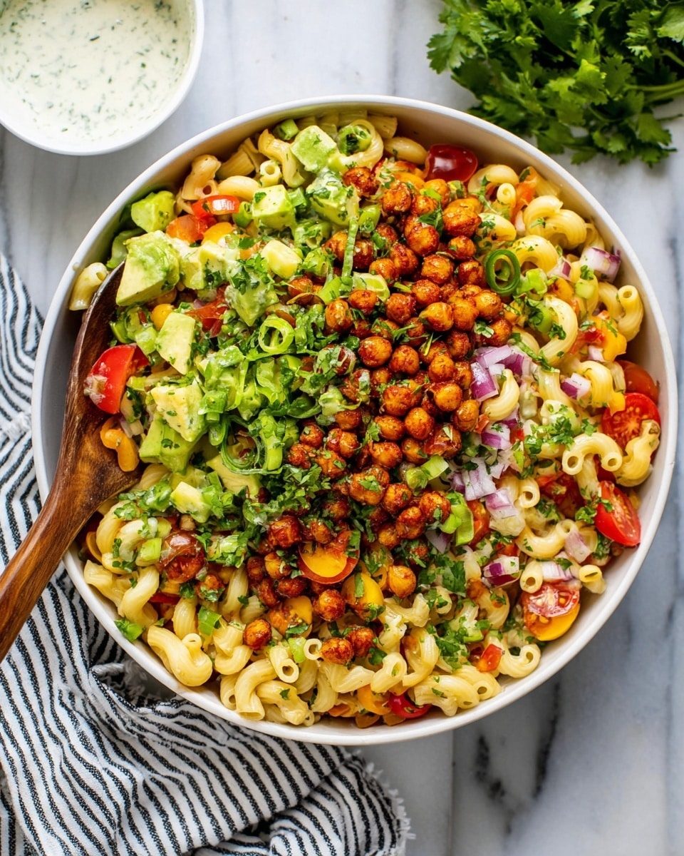 A large white bowl is filled with three layers: the base layer is light yellow pasta curved in small tubes mixed with tiny pieces of red and yellow bell peppers, cherry tomato halves, and finely chopped red onions. On top of this, there is a layer of roasted chickpeas that are golden brown and crispy. The final layer includes green slices of avocado, chopped green onions, and chopped fresh cilantro scattered all over. A wooden spoon is placed inside the bowl on the left side. The bowl is set on a white marbled surface with a striped cloth nearby, and a small white bowl of white dressing is visible in the upper left corner. photo taken with an iphone --ar 4:5 --v 7