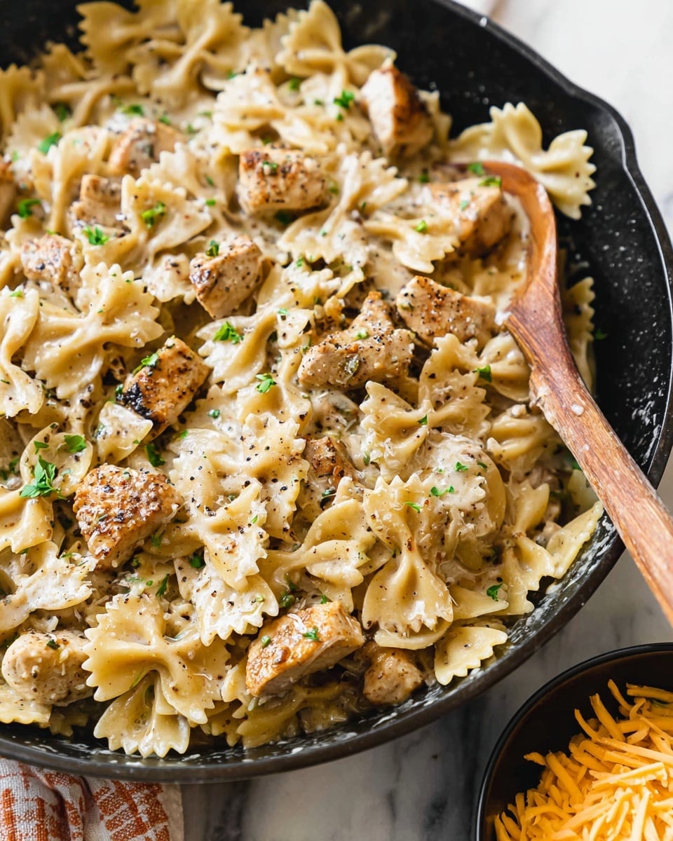 A close-up view of a black pan filled with farfalle pasta layered with chunks of golden-brown cooked chicken, coated in a creamy light beige sauce speckled with black pepper and garnished with small green herb pieces scattered unevenly across the top. A wooden spoon rests in the pan, partially submerged in the pasta. The pan sits on a white marbled surface with a glimpse of a black bowl with an orange rim filled with shredded cheese in the bottom right corner. photo taken with an iphone --ar 4:5 --v 7