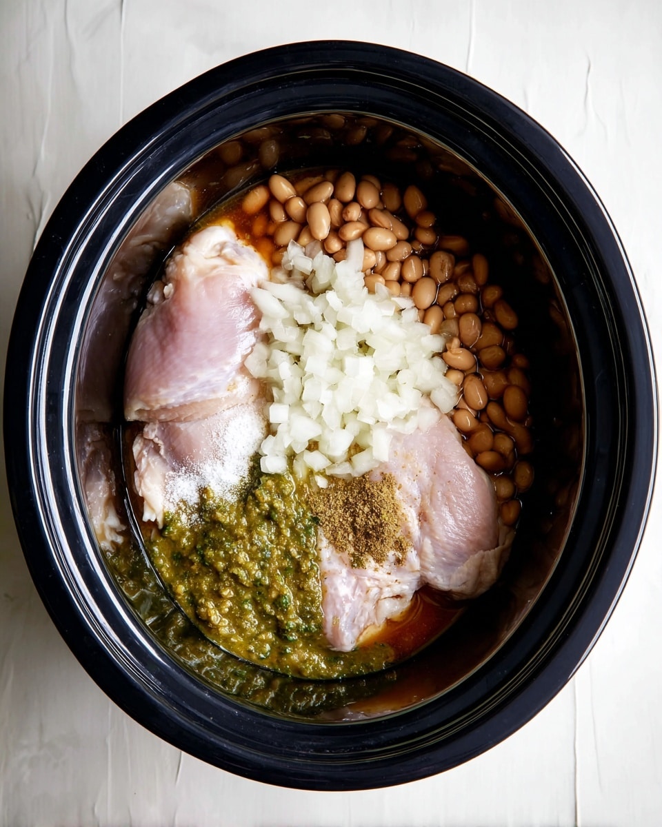 Inside a black slow cooker, there are several ingredients arranged in layers. At the bottom, there are two raw light pink chicken pieces, one slightly overlapping the other. On top of the chicken, there are chopped white onions scattered in the middle, light brown beans clustered on the right side, and a mix of green salsa spread in patches near the onions and beans. Light-colored spices are sprinkled over the onions and beans, with a small pile of white salt visible. The slow cooker sits on a white marbled surface. photo taken with an iphone --ar 4:5 --v 7