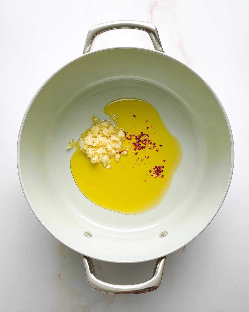 A white cooking pot with two metal handles sits on a white marbled surface. Inside the pot, there is a small pool of yellow olive oil, with a beige clump of minced garlic near the top left of the oil, and a small sprinkle of red chili flakes near the bottom right of the oil. The pot's inside surface is smooth and light gray, creating a clean and simple setting for the ingredients. photo taken with an iphone --ar 4:5 --v 7