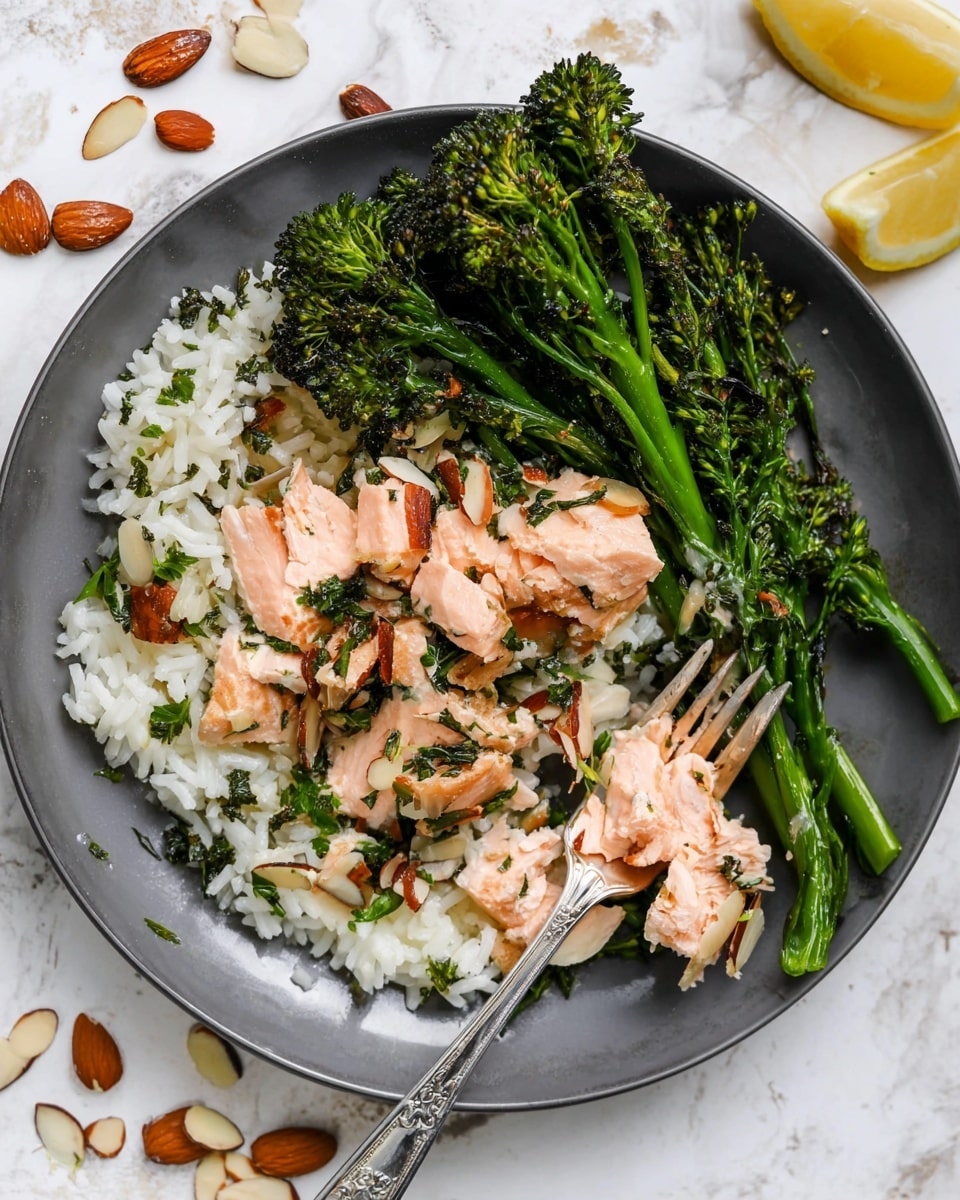 A white plate holds three main layers: at the bottom, there is a bed of white rice mixed with green herbs; on top of this, chunks of pink cooked salmon are scattered, mixed with sliced brown almonds; on one side, bright green charred broccolini is placed in a small bunch. A silver fork rests on the edge of the plate. The plate is set on a white marbled surface with scattered almond slices and two lemon pieces nearby. Photo taken with an iphone --ar 4:5 --v 7