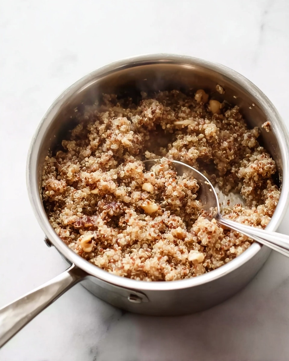A close-up view of a shiny silver pot filled with cooked quinoa mixed with small pieces of a light brown ingredient, possibly nuts or vegetables, giving the dish a lumpy texture. The quinoa mixture has a soft, cooked look with many tiny white spiral shapes visible. A silver spoon rests inside the pot, partially submerged in the quinoa mix, and the whole scene sits on a white marbled surface. Steam rises gently from the pot, indicating the food is hot. photo taken with an iphone --ar 4:5 --v 7