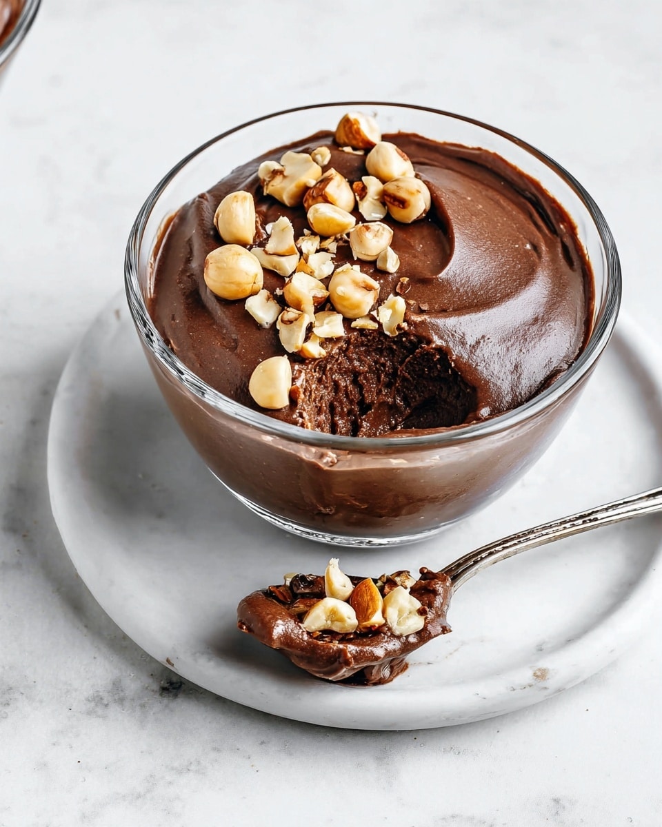 The image shows a small clear glass bowl filled with a smooth, dark chocolate mousse that has a rich, creamy texture. On top of the mousse, there is a line of roughly chopped hazelnuts scattered, adding a light brown color contrast and a crunchy texture. The bowl is placed on a simple white plate with a white marbled surface underneath. Next to the bowl, a silver spoon holds a dollop of the same chocolate mousse with a few hazelnuts stuck to it. The overall look is clean and inviting, focusing on the thick, shiny mousse and nut topping. photo taken with an iphone --ar 4:5 --v 7