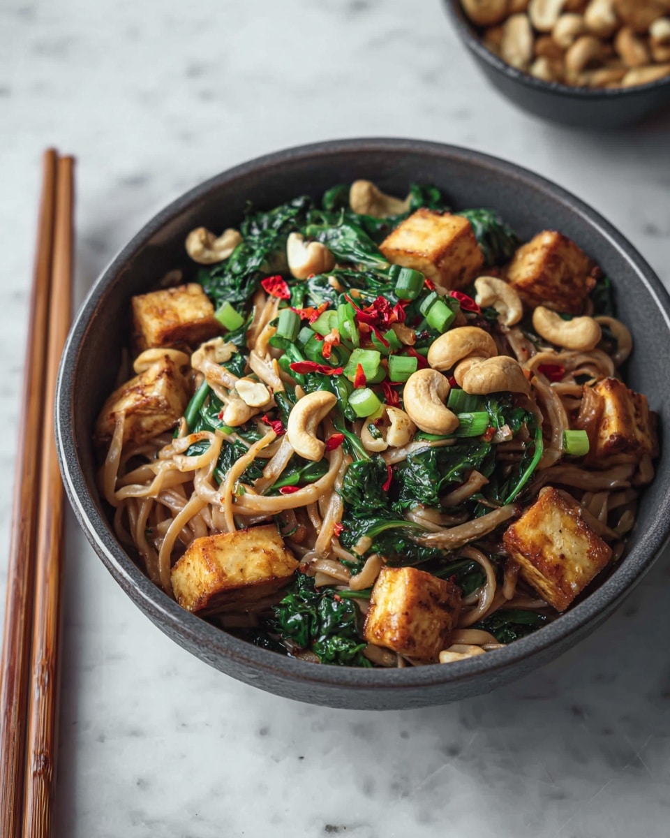 A bowl filled with a noodle dish is shown on a white marbled surface, with a pair of wooden chopsticks on the left. The bowl is dark gray and round. Inside, thick, light brown noodles form the base layer, mixed with deep green leafy vegetables. Scattered on top are golden-brown cubed tofu pieces and whole roasted cashews, adding a crunchy texture. Small red chili flakes and chopped green onions are sprinkled over the dish, giving bright pops of red and green color. Photo taken with an iphone --ar 4:5 --v 7