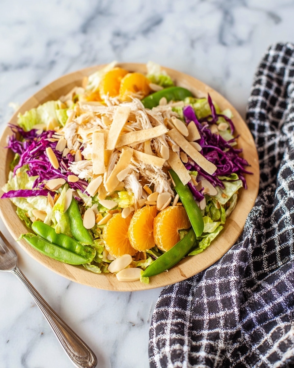 The image shows a colorful salad served on a light wooden plate placed on a white marbled surface. The salad has several layers starting with a base of green lettuce and bright purple cabbage, topped with slices of mandarin orange and snap peas. There is shredded chicken layered above the vegetables, followed by crunchy light beige strips placed on the top. Scattered sunflower seeds and almond slices add texture and variety. A black and white checkered cloth is partially seen on the right side of the image, and a silver fork is placed on the bottom left. photo taken with an iphone --ar 4:5 --v 7