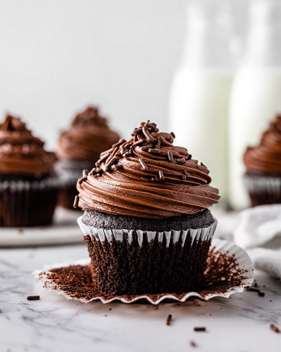 A close-up of a rich chocolate cupcake with one layer of dark, moist cake at the bottom, topped by a thick swirl of smooth, shiny chocolate frosting in dark brown, decorated with small chocolate sprinkles. The cupcake liner is open at the bottom and the cupcake sits on a white marbled surface. In the blurred background, there are three more chocolate cupcakes standing on the same white marbled surface and two tall glass bottles of milk. Photo taken with an iphone --ar 4:5 --v 7