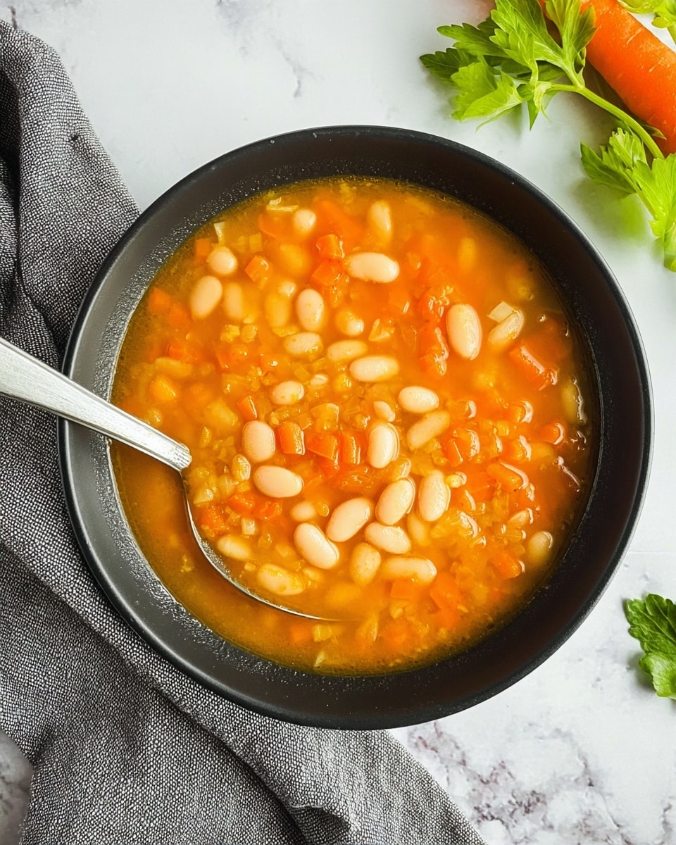 A close-up of a black bowl filled with chunky orange soup, showing at least three layers: a clear orange broth base, scattered white beans as the middle layer, and small diced orange carrot pieces mixed evenly throughout the soup as the top layer. A silver spoon rests inside the bowl on the left side. The bowl sits on a white marbled surface with a grey cloth napkin draped beside it on the left, fresh green celery leaves at the top right corner, and part of an orange carrot visible on the right edge. Photo taken with an iphone --ar 4:5 --v 7