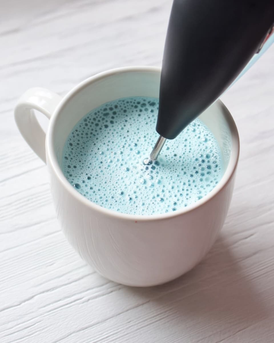 A white ceramic mug filled nearly to the top with a light blue foamy liquid, with many small bubbles covering the surface. A black handheld electric frother is submerged in the liquid from above, creating the foam. The mug sits on a white marbled textured surface, and a woman's hand is holding the frother just out of view. The image is bright and softly lit. photo taken with an iphone --ar 4:5 --v 7