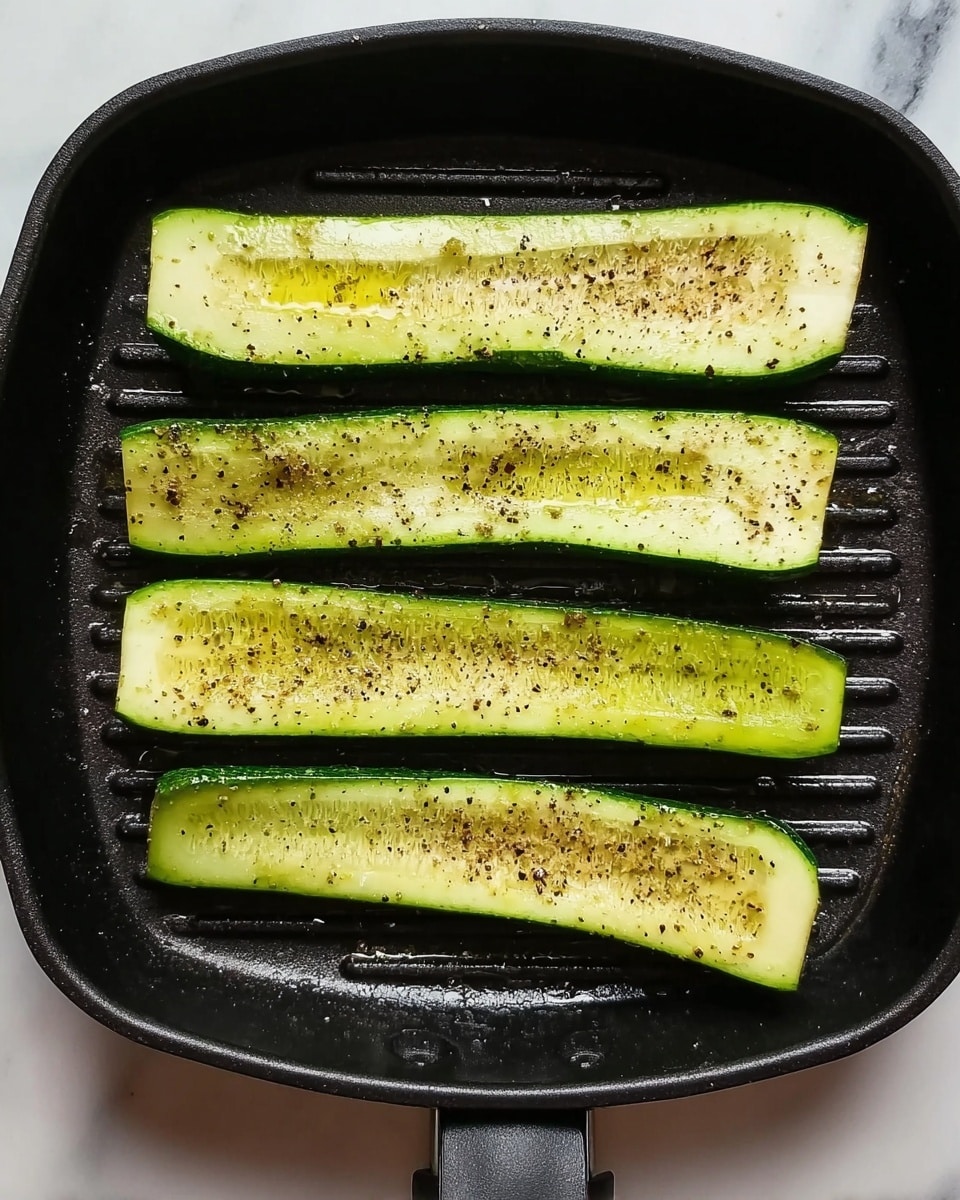 The image shows a black grill pan with five long slices of green zucchini placed side by side inside it. Each zucchini slice is seasoned with black pepper and some oil, giving the bright green vegetables a slightly shiny texture. The grill pan sits on a white marbled surface, and the focus is on the simple, fresh look of the vegetables in the dark pan. Photo taken with an iphone --ar 4:5 --v 7