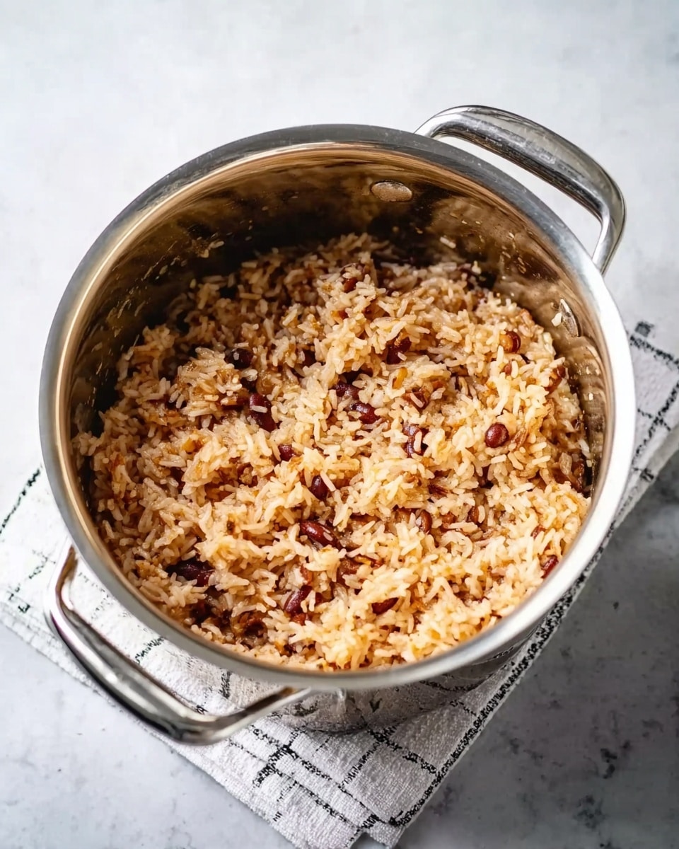 A stainless steel pot filled with cooked rice mixed with finely chopped red beans and small brown bits. The rice looks soft and creamy, with a mix of light brown and reddish colors evenly spread throughout. The pot has two metal handles and sits on a folded white and black checkered cloth, all placed on a white marbled surface. The lighting is soft and natural, highlighting the texture of the rice and beans clearly. photo taken with an iphone --ar 4:5 --v 7