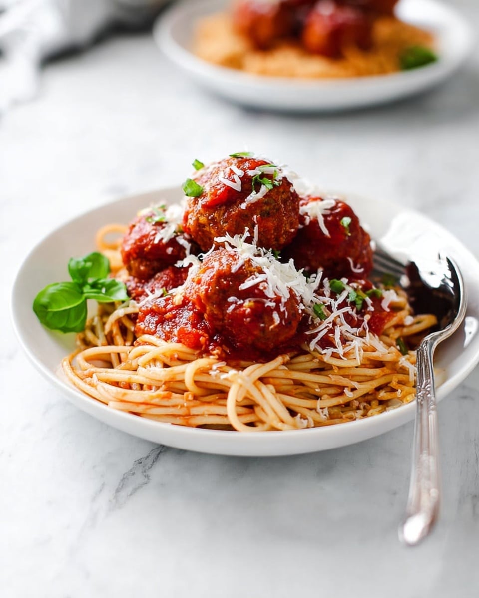 A white plate holds a neat round bed of light brown spaghetti noodles at the bottom. On top of the noodles, there are four large reddish-brown meatballs covered in bright red tomato sauce with small chunks. The meatballs and sauce are lightly sprinkled with white shredded cheese, and a small green basil leaf sits on the side for garnish. A silver fork and spoon rest together on the right side of the plate, touching the noodles. The plate sits on a white marbled textured surface with a blurry second plate of the same dish in the background. Photo taken with an iphone --ar 4:5 --v 7
