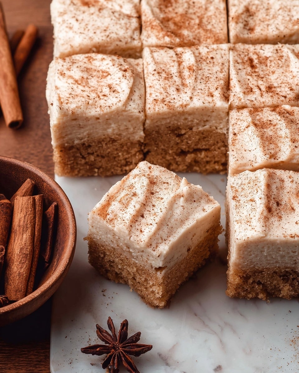 The image shows a dessert made of square pieces arranged closely together, with one piece pulled slightly away to reveal the inside. There are two main layers: the bottom layer is a light brown, moist, and soft cake with a crumbly texture, while the top layer is a thick, creamy frosting in a pale beige color, sprinkled evenly with a fine dusting of cinnamon or spice, adding a textured look. The cake pieces rest on a white marbled surface, and to the left, there is a wooden bowl containing cinnamon sticks and star anise, adding a warm, rustic feeling to the scene. photo taken with an iphone --ar 4:5 --v 7