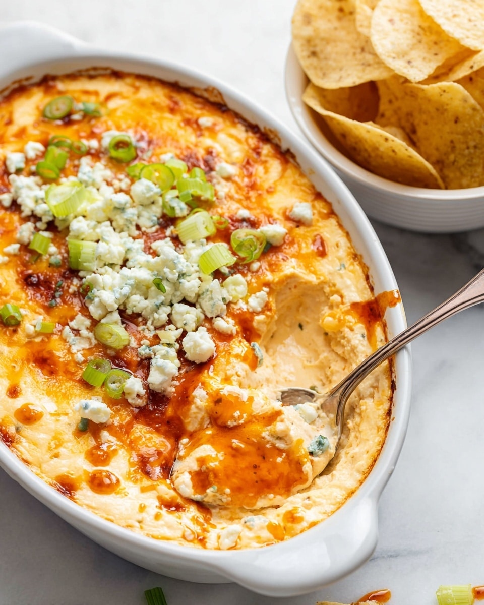 A white oval dish holds a layered dip with three visible layers: the bottom layer is creamy and pale yellow, the middle layer is a mix of melted, golden-brown cheese with some bubbly browned spots, and the top layer is dotted with crumbled white cheese and chopped green onions scattered across. There is some orange sauce drizzled over the top. A silver spoon rests inside the dish, scooping out a portion of the dip to show its creamy texture. To the top right, there is a white bowl filled with triangular pale yellow tortilla chips placed on a white marbled surface photo taken with an iphone --ar 4:5 --v 7
