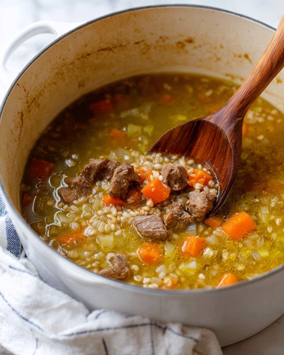 The image shows a bowl of beef and barley soup with visible layers of ingredients. At the bottom is a clear, light brown broth. On top, there are chunky pieces of cooked brown beef and small diced orange carrots evenly spread. The barley grains, pale beige and plump, are mixed with the carrots and beef in the soup. Fresh green herb bits are sprinkled all over, adding a touch of color. The soup is in a white bowl with a speckled brown rim, and a metal spoon is resting inside. The bowl is placed on a white marbled surface with some green herbs and part of a striped cloth nearby. Photo taken with an iphone --ar 4:5 --v 7