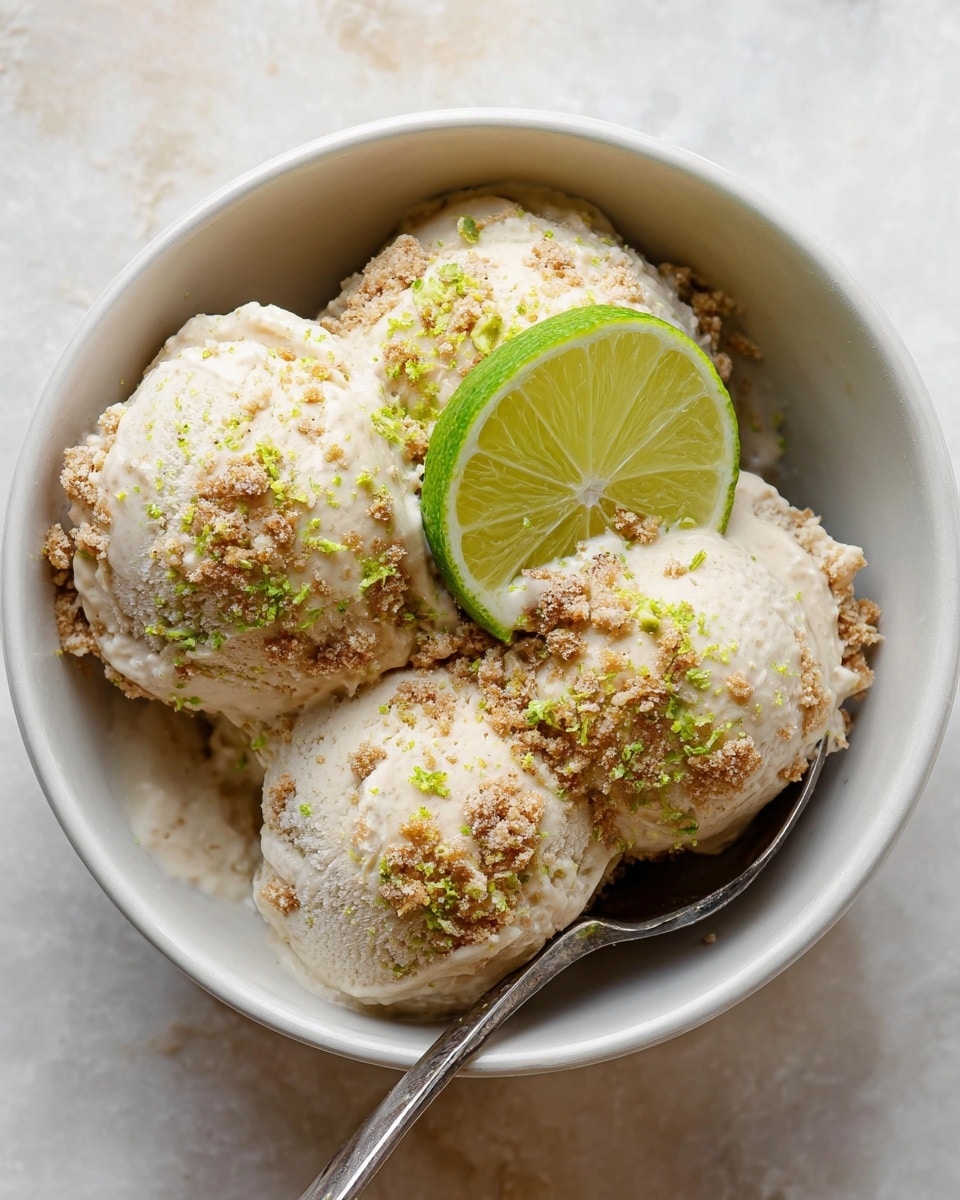 A white bowl holds three scoops of light beige ice cream with a slightly crumbly texture and small brown bits mixed in. The ice cream scoops are sprinkled with fine green zest and brown crumble, giving a textured look. On top of the middle scoop, there is a bright green lime wedge with visible segments. A silver spoon rests inside the bowl against the side. The bowl sits on a white marbled surface. Photo taken with an iphone --ar 4:5 --v 7
