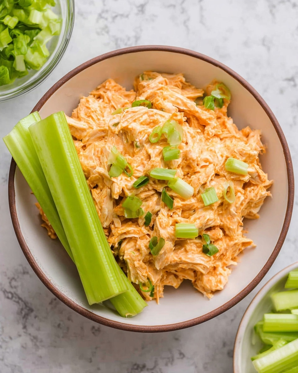 A bowl filled with a creamy mix of shredded chicken coated in an orange sauce, topped with small pieces of green onion scattered on top. Two large bright green celery sticks rest upright on the side inside the bowl. The bowl is white with a thin brown rim, placed on a white marbled background. Part of a glass bowl with more chopped green onions and a white bowl of celery pieces are visible around the main bowl. Photo taken with an iphone --ar 4:5 --v 7