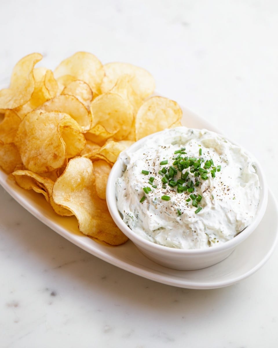 A white oval plate with a white round bowl on the left side filled with creamy white dip, which has a thick, smooth texture with small green chive pieces mixed in. The dip is topped with finely chopped green chives and a sprinkle of black pepper. To the right of the bowl, a pile of light golden potato chips is spread out on the plate. The whole setup is placed on a white marbled surface. photo taken with an iphone --ar 4:5 --v 7