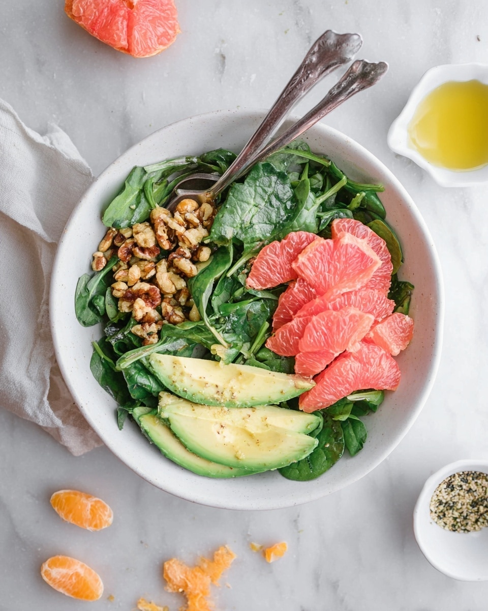 A white bowl sits on a white marbled surface, filled with a fresh salad composed of three main layers. The bottom layer is a bed of dark green spinach leaves with some smaller leafy greens mixed in, giving a rich textured look. On the left side above the greens, there is a cluster of golden brown roasted nuts, adding a rough, crunchy texture. On the right side, several bright pink grapefruit segments with a juicy, slightly shiny surface are arranged neatly in a small pile. Above the grapefruit, there are three avocado slices with pale green creamy flesh, slightly curved and smooth. A silver spoon and fork rest inside the bowl, leaning against each other near the top edge. Scattered around the bowl on the marbled surface are some small bits of grapefruit, some orange slices, a small white bowl with seeds, and a small cup of olive oil. photo taken with an iphone --ar 4:5 --v 7