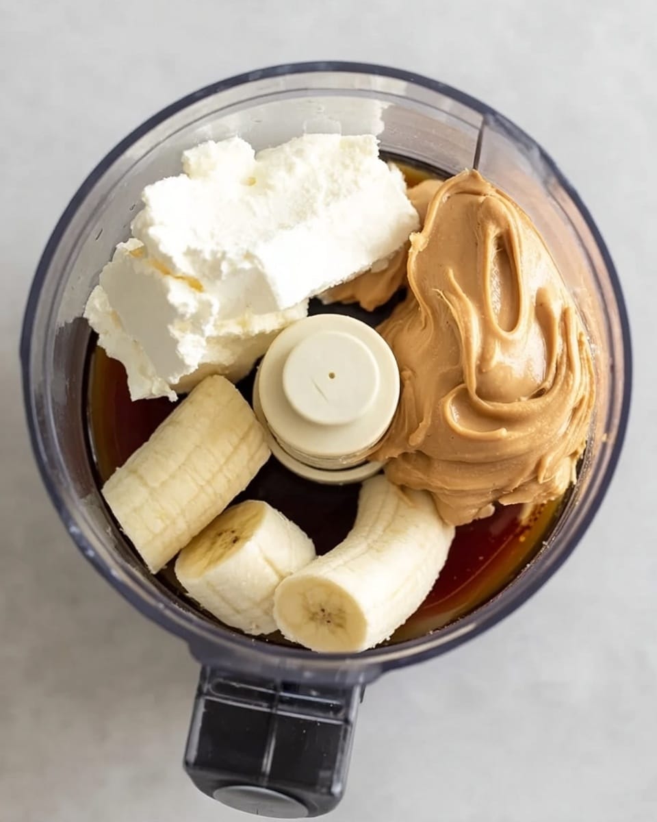 A round clear food processor bowl sits on a white marbled surface, containing four main ingredients separated but close together. On the left, there is a large chunk of cream cheese, white and smooth with soft edges. Next to it on the right, there is a big scoop of peanut butter, light brown and creamy with visible swirls. Below these two, a peeled banana is broken into three pieces, light yellow in color with a soft texture. The base of the bowl holds a dark amber liquid, likely syrup, surrounding the ingredients and filling the bottom of the processor. The image captures the clear plastic sides and center blade of the food processor, showing all the details inside. Photo taken with an iphone --ar 4:5 --v 7
