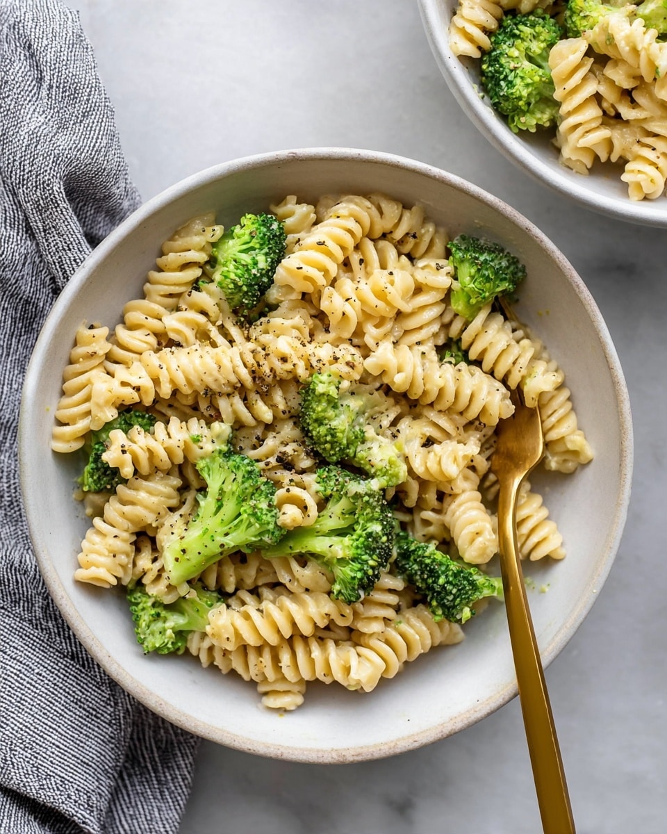 The image shows a white bowl filled with short spiral pasta mixed evenly with bright green broccoli florets. The pasta is creamy light yellow and the broccoli adds a fresh green color, creating a balanced look. The dish is sprinkled with some black pepper mostly on the pasta surface. A gold fork is placed inside the bowl, resting on the pasta near the bottom right side. The bowl sits on a white marbled surface, and a part of a grey striped cloth is visible at the bottom left corner. Another white bowl with the same food is partially visible in the top right corner. Photo taken with an iphone --ar 4:5 --v 7