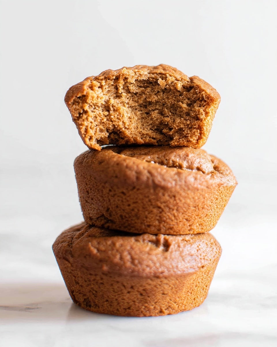 Three small, round brown muffins stacked on top of each other on a white marbled surface with a plain white background; the top muffin is broken in half and sits on the second muffin, showing its moist and dense inside texture with a slightly cracked top crust; the muffins have a soft, slightly rough surface with a warm golden-brown color; photo taken with an iphone --ar 4:5 --v 7