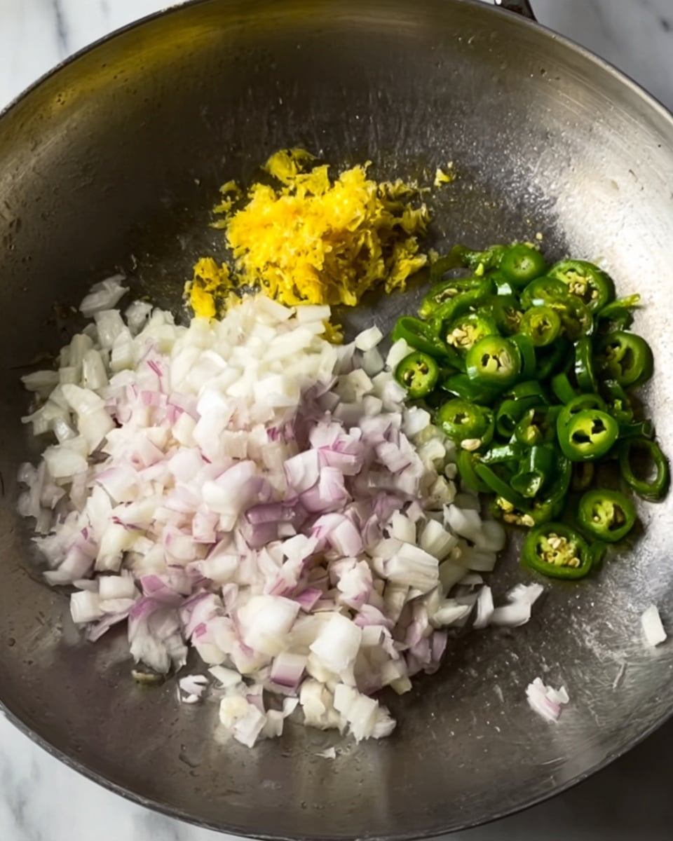 The image shows a close-up of a metal pan with three separate piles of chopped ingredients arranged inside it. At the bottom center, there is a pile of finely chopped white and light purple onions with a soft texture. To the left, near the edge of the pan, there is a small heap of finely minced yellow ginger. On the right side, there are sliced green chili peppers with a fresh, shiny appearance. The pan is placed on a white marbled surface. Photo taken with an iphone --ar 4:5 --v 7