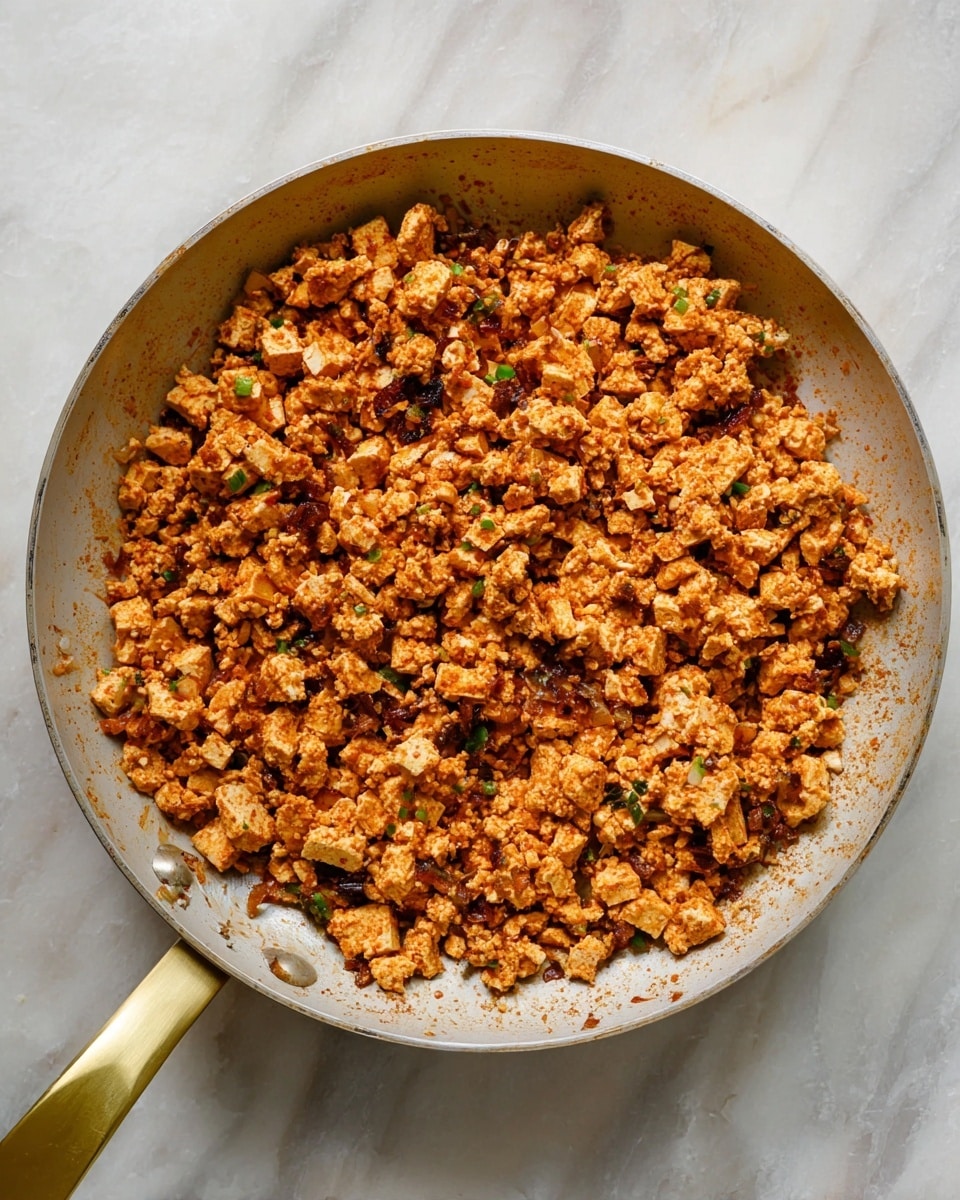 A white pan with a shiny gold handle holds a mix of cooked crumbled tofu and diced onions, all coated in a soft reddish-brown seasoning. The tofu pieces are small and uneven, mixed with lightly browned onion bits that add texture. The dish fills the pan evenly, showing a mix of crumbly and chunky textures, with some green hints scattered throughout. The background is a white marbled surface. photo taken with an iphone --ar 4:5 --v 7
