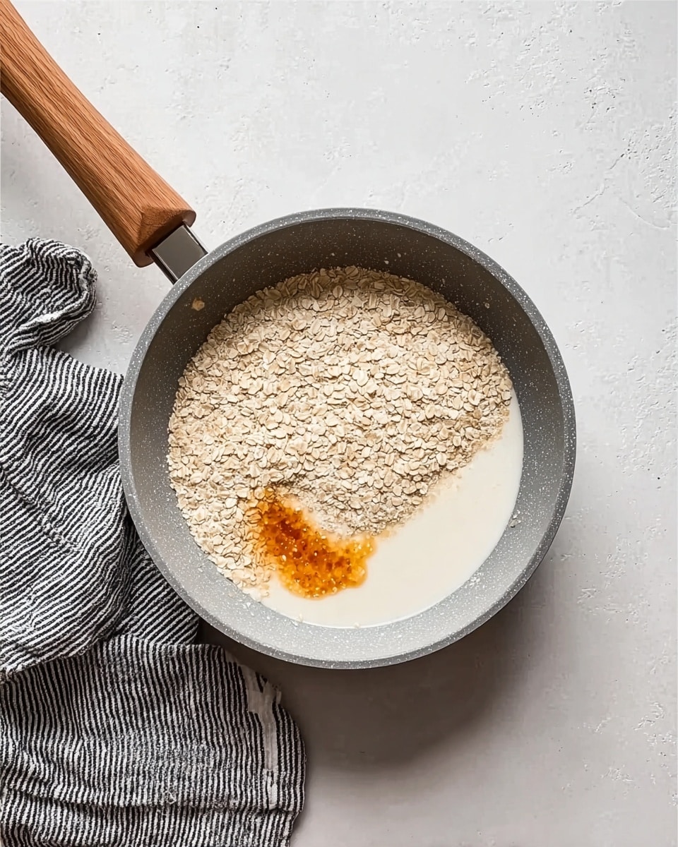 A grey frying pan with a wooden handle sits on a white marbled surface next to a folded black and white striped cloth. Inside the pan, there is a layer of light beige oats spread evenly, a small pool of white milk creating a smooth area on one side, and a dollop of orange syrup or liquid on the milk. The textures contrast softly with the rough oats, smooth milk, and shiny syrup. Photo taken with an iphone --ar 4:5 --v 7