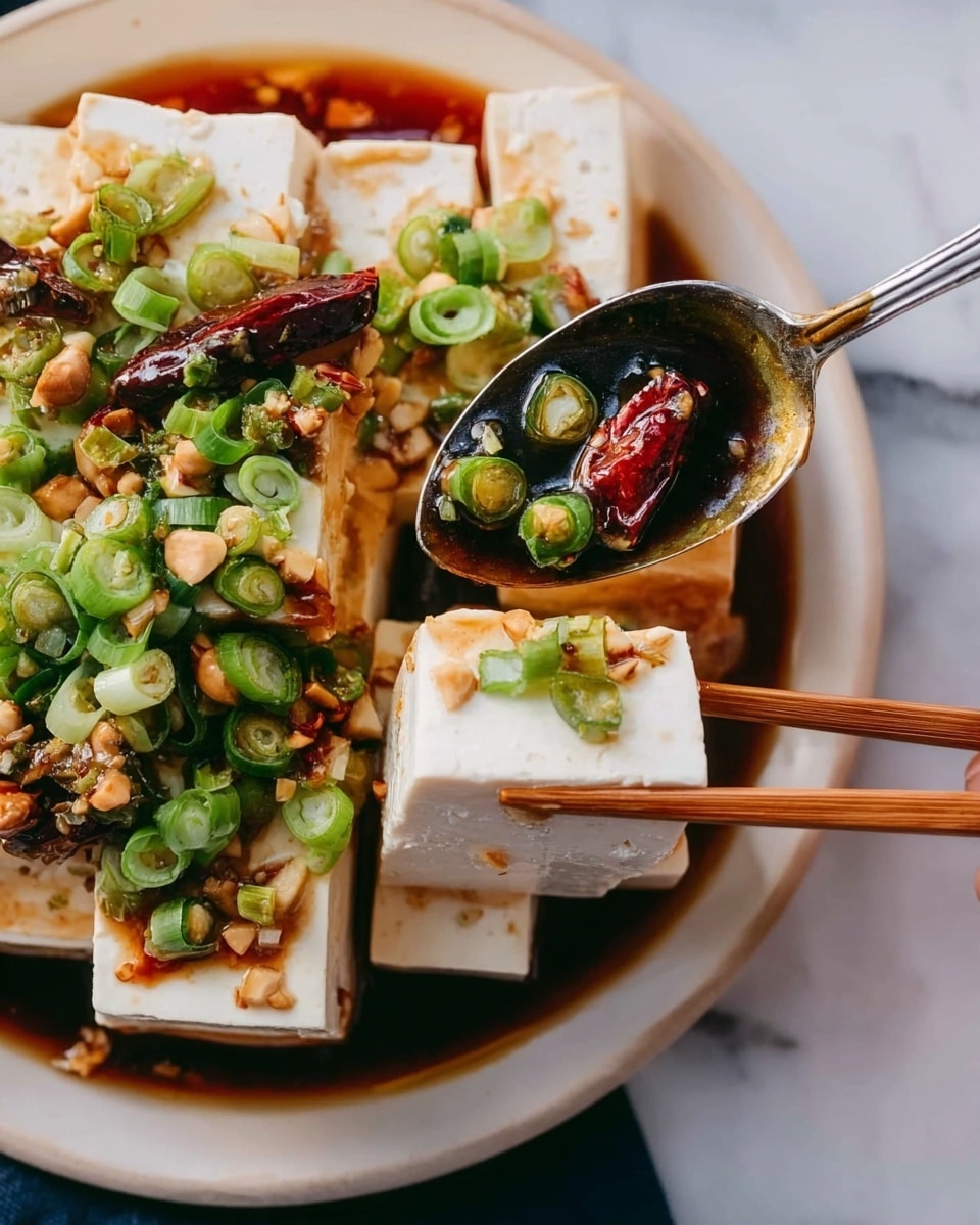 The image shows a close-up of soft tofu blocks arranged on a white plate, topped with a dark brown sauce that looks oily and slightly thick. On top of the tofu, there are bright green sliced spring onions scattered generously, and a few whole dried red chili peppers add a pop of color. Some chopped nuts or crunchy bits are sprinkled over the tofu and sauce, providing texture contrast. A piece of tofu is being held by a pair of wooden chopsticks near the bottom right side, and a woman's hand holds a silver spoon scooping up tofu from the left side of the plate. The background is a white marbled surface. photo taken with an iphone --ar 4:5 --v 7