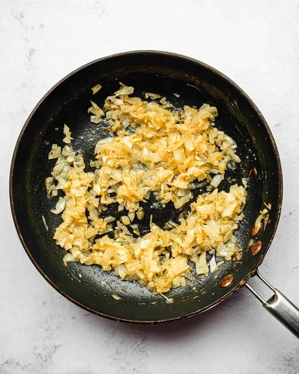 A black pan filled with a small amount of cooked onions that are soft and light golden in color, spread unevenly across the pan's surface. The onions have a slightly shiny texture, showing they are cooked with some oil or butter. The pan's black interior contrasts with the pale golden onions. The pan rests on a white marbled surface, with the handle visible on the right side. There are some marks and slight burns on the pan edge, showing it has been used for cooking. photo taken with an iphone --ar 4:5 --v 7
