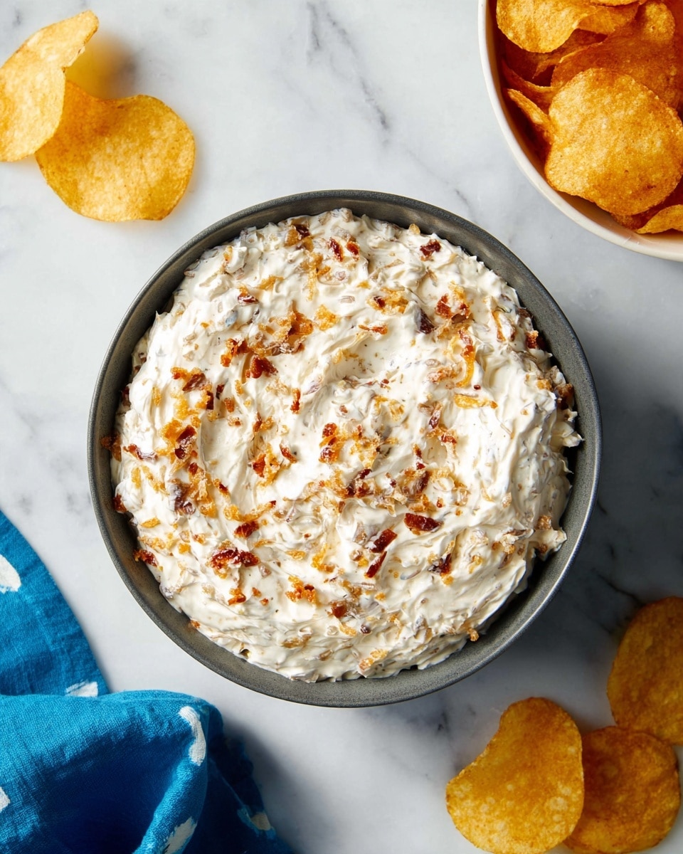 A round bowl filled with creamy white dip mixed with bits of golden brown and slightly darker crispy pieces, creating a textured surface with specks of color throughout. The bowl is simple and dark gray, placed on a white marbled surface. To the right, there is another white bowl, filled with golden, crispy potato chips stacked loosely. Three potato chips lie scattered on the surface near the bowls. A blue cloth with light white spots is softly draped at the bottom left corner of the image, adding a splash of color. Photo taken with an iphone --ar 4:5 --v 7