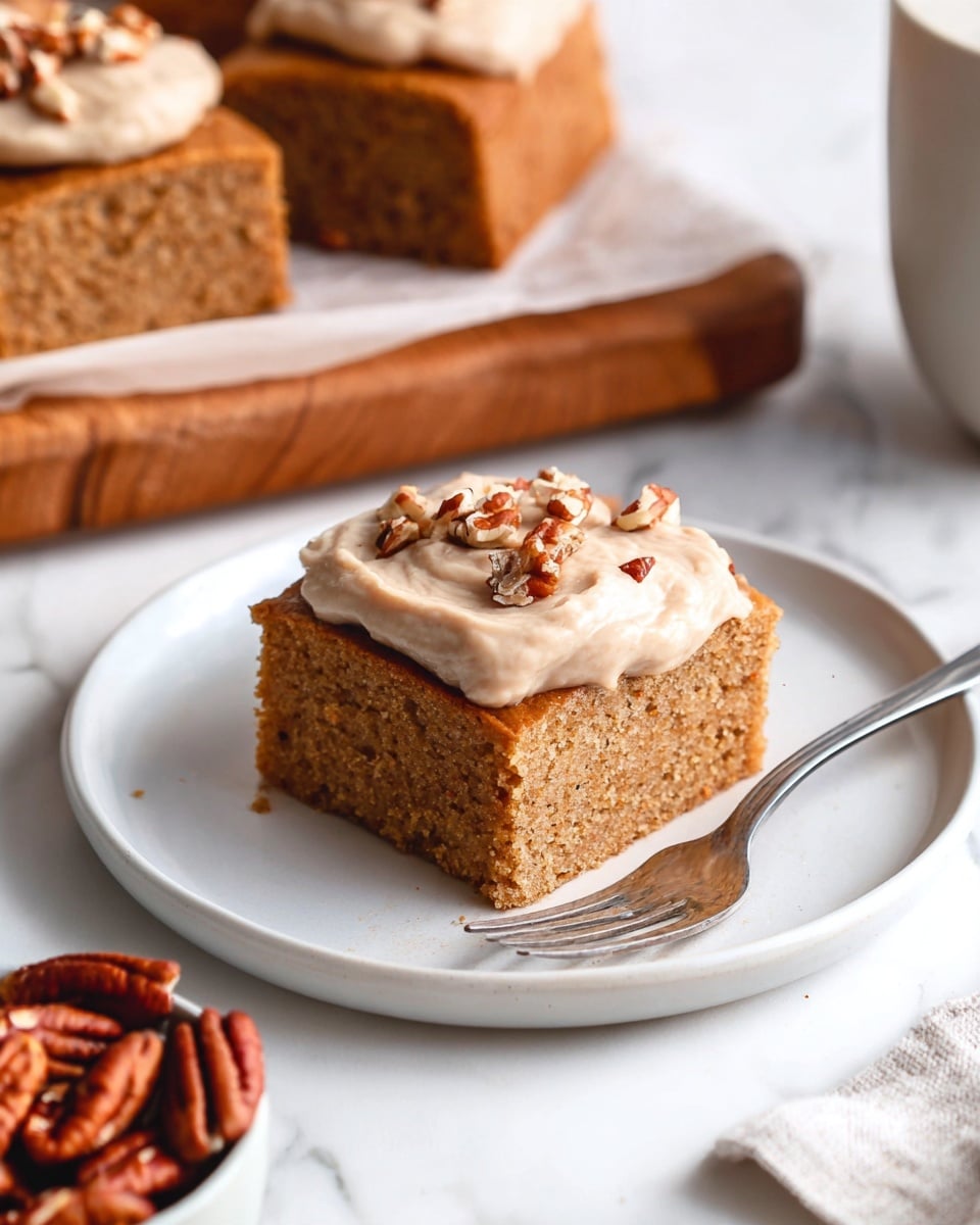A square piece of light brown cake with a soft texture is placed in the center of a white plate. On top of the cake, there is a dollop of light beige frosting with a smooth texture, sprinkled with small pieces of chopped pecans. A silver fork lies on the right side of the plate, angled slightly towards the cake. In the background, additional cake pieces with a similar light brown color sit on a wooden board lined with white parchment paper, and on the left bottom corner, a small bowl filled with whole pecans is visible. The surface beneath everything has a white marbled texture. Photo taken with an iphone --ar 4:5 --v 7