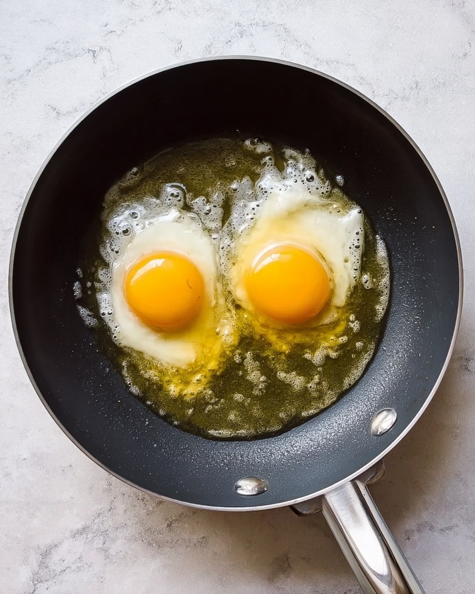 A black frying pan with a shiny silver handle contains two raw eggs cooking in oil. The eggs are side by side, with bright yellow yolks and clear, slightly white egg whites spreading unevenly in the warm oil. The pan sits on a white marbled surface that softly reflects a bit of light. The oil looks warm and slightly bubbling around the edges of the eggs. photo taken with an iphone --ar 4:5 --v 7